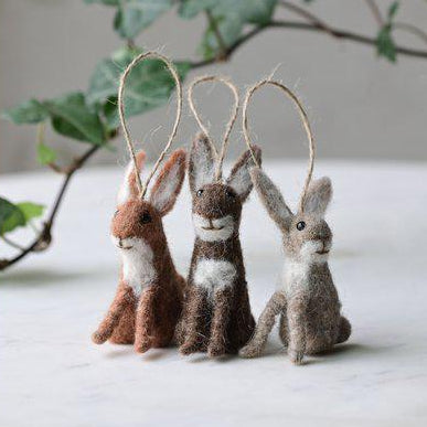 Three felted rabbit ornaments on a white surface with green leaves in the background