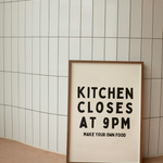 Framed poster with text 'Kitchen Closes at 9PM Make Your Own Food' against a tiled wall.