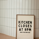 Framed poster with text 'Kitchen Closes at 9PM Make Your Own Food' against a tiled wall.