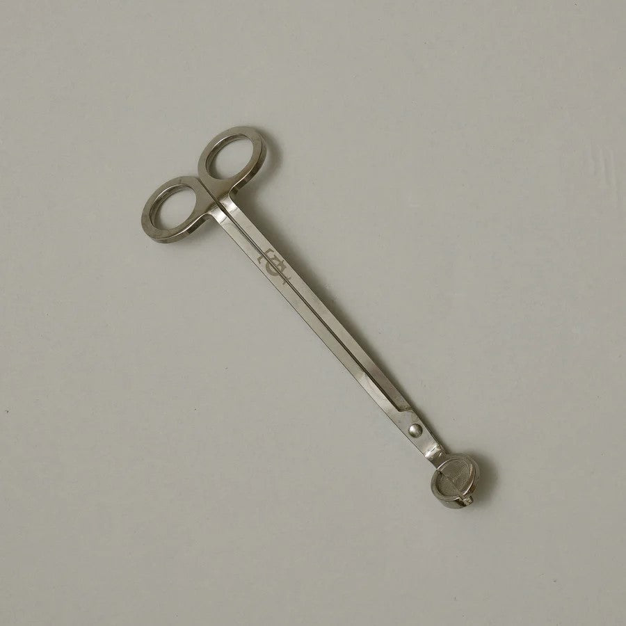 Stainless steel wick trimmer with a looped end and the True Grace emblem engraved on the handle