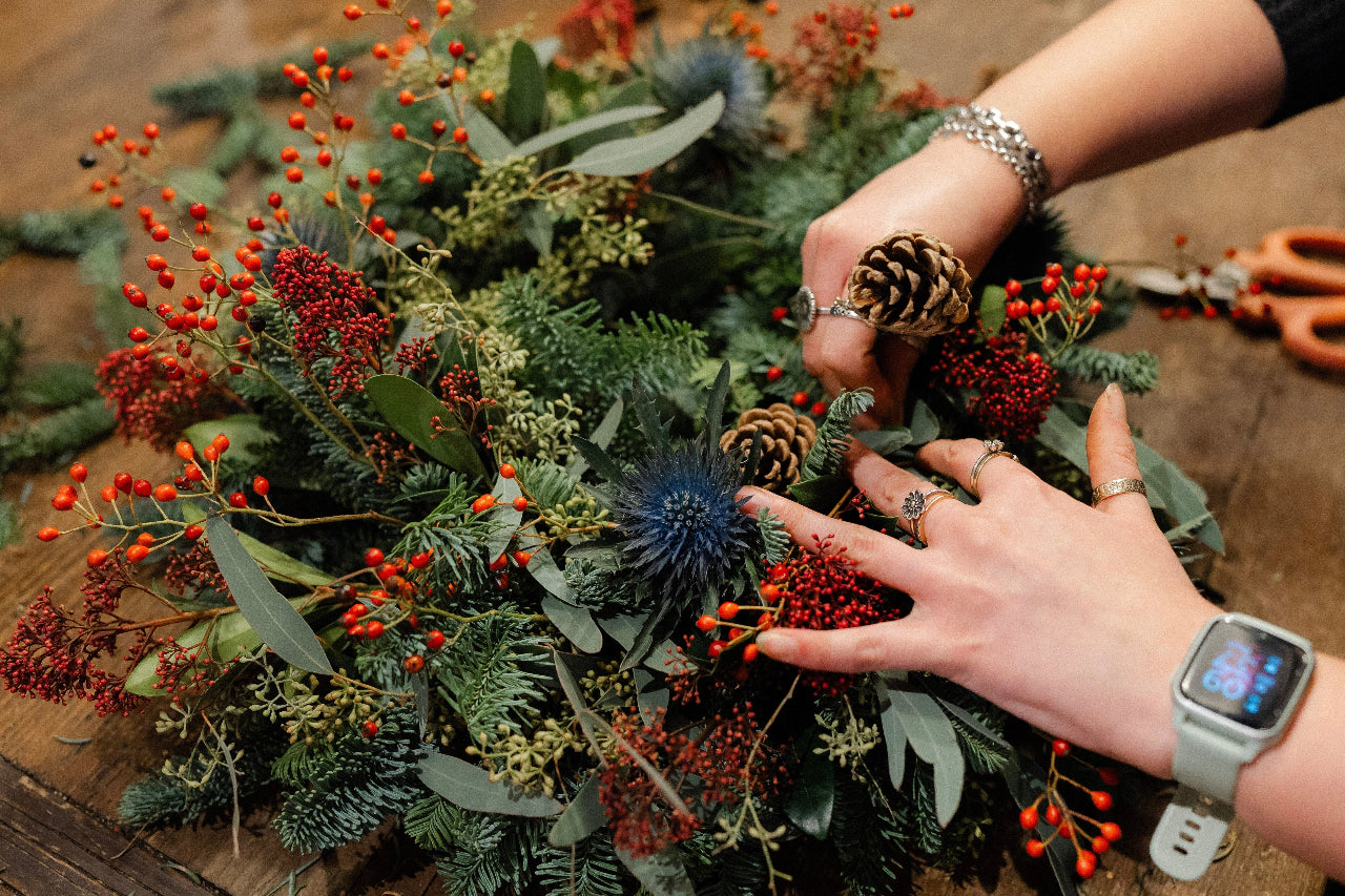Person arranging a festive wreath with pine cones and berries on a wooden surface.