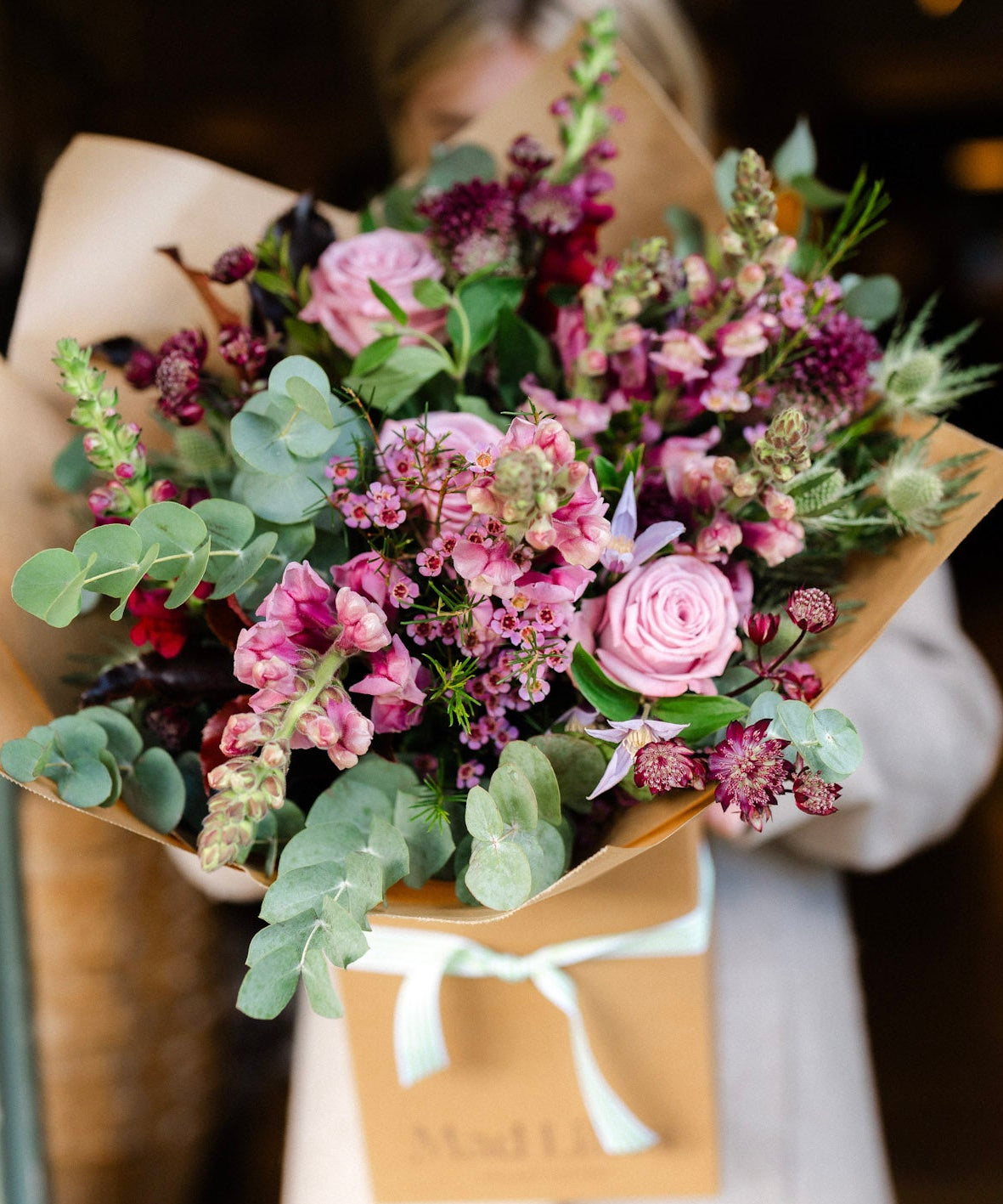 Person holding a bouquet of flowers wrapped in brown paper with a white ribbon.