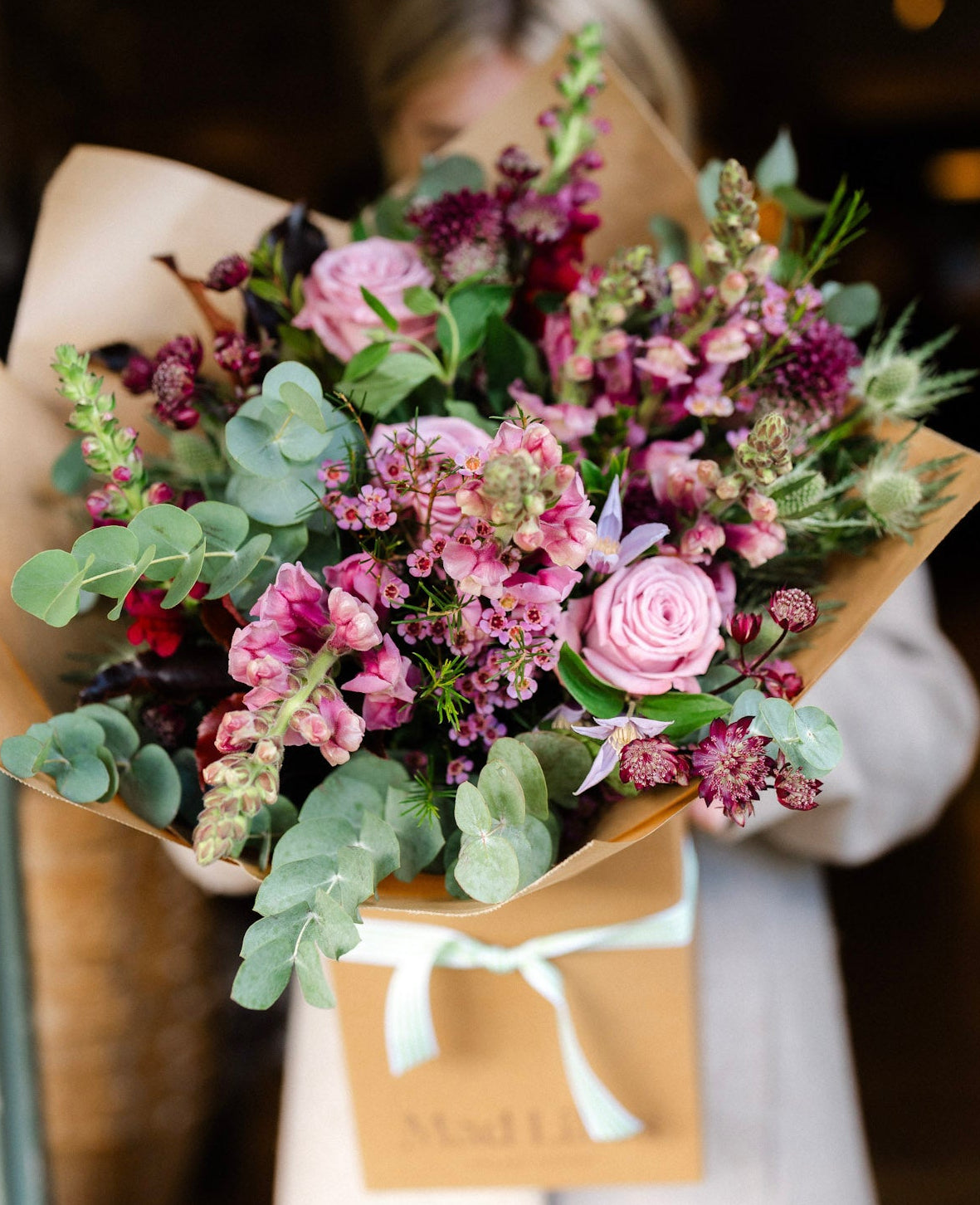 Person holding a bouquet of flowers wrapped in brown paper with a white ribbon.
