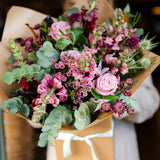Person holding a bouquet of flowers wrapped in brown paper with a white ribbon.