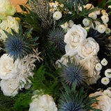 A rustic floral arrangement featuring white and pale green flowers, blue thistle accents, and green foliage, displayed in a wooden box on a rustic wooden table in a styled indoor setting.
