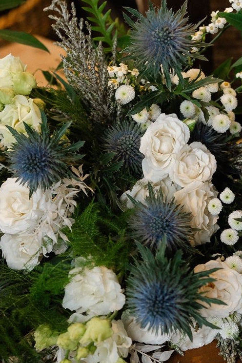 A rustic floral arrangement featuring white and pale green flowers, blue thistle accents, and green foliage, displayed in a wooden box on a rustic wooden table in a styled indoor setting.