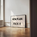 Framed poster with text 'NEW PLAN: FUCK IT' in a room with wooden floor and white walls.