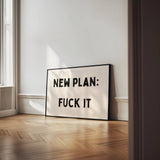 Framed poster with text 'NEW PLAN: FUCK IT' in a room with wooden floor and white walls.