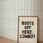 Framed poster with 'Boots Off Here, Cowboy' text against a tiled wall.