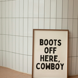 Framed poster with 'Boots Off Here, Cowboy' text against a tiled wall.