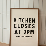 Framed poster with text 'Kitchen Closes at 9PM Make Your Own Food' against a tiled wall.