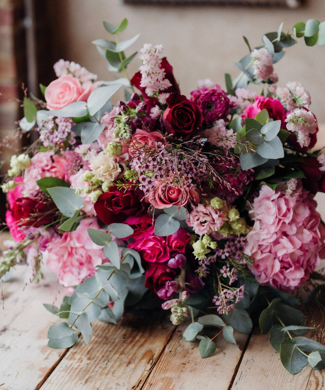 A luxurious floral arrangement featuring a marshmallow pink and rich marsala red colour palette, with a variety of flowers including roses, hydrangeas, snapdragons, lisianthus, and seasonal foliage