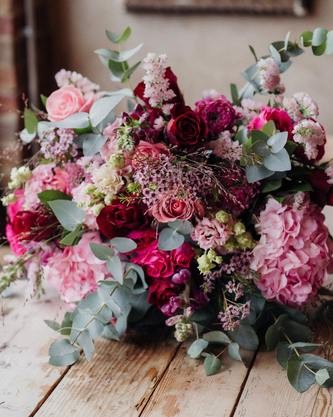 A luxurious floral arrangement featuring a marshmallow pink and rich marsala red colour palette, with a variety of flowers including roses, hydrangeas, snapdragons, lisianthus, and seasonal foliage