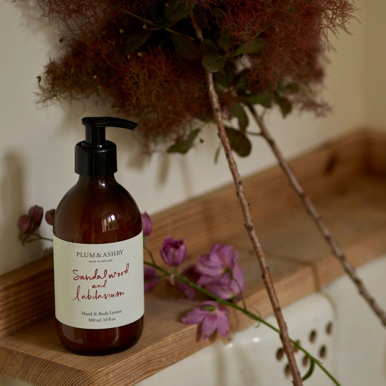 A pump bottle of Plum & Ashby Sandalwood & Labdanum hand and body lotion placed on a wooden surface with dried flowers and a lit candle in the background, giving a warm, cozy ambiance.