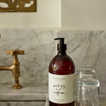 1 Litre bottle of Plum & Ashby Wild Fig & Saffron Hand & Body Wash on a kitchen countertop with a marble backdrop and a gold-patterned napkin holder in the background.