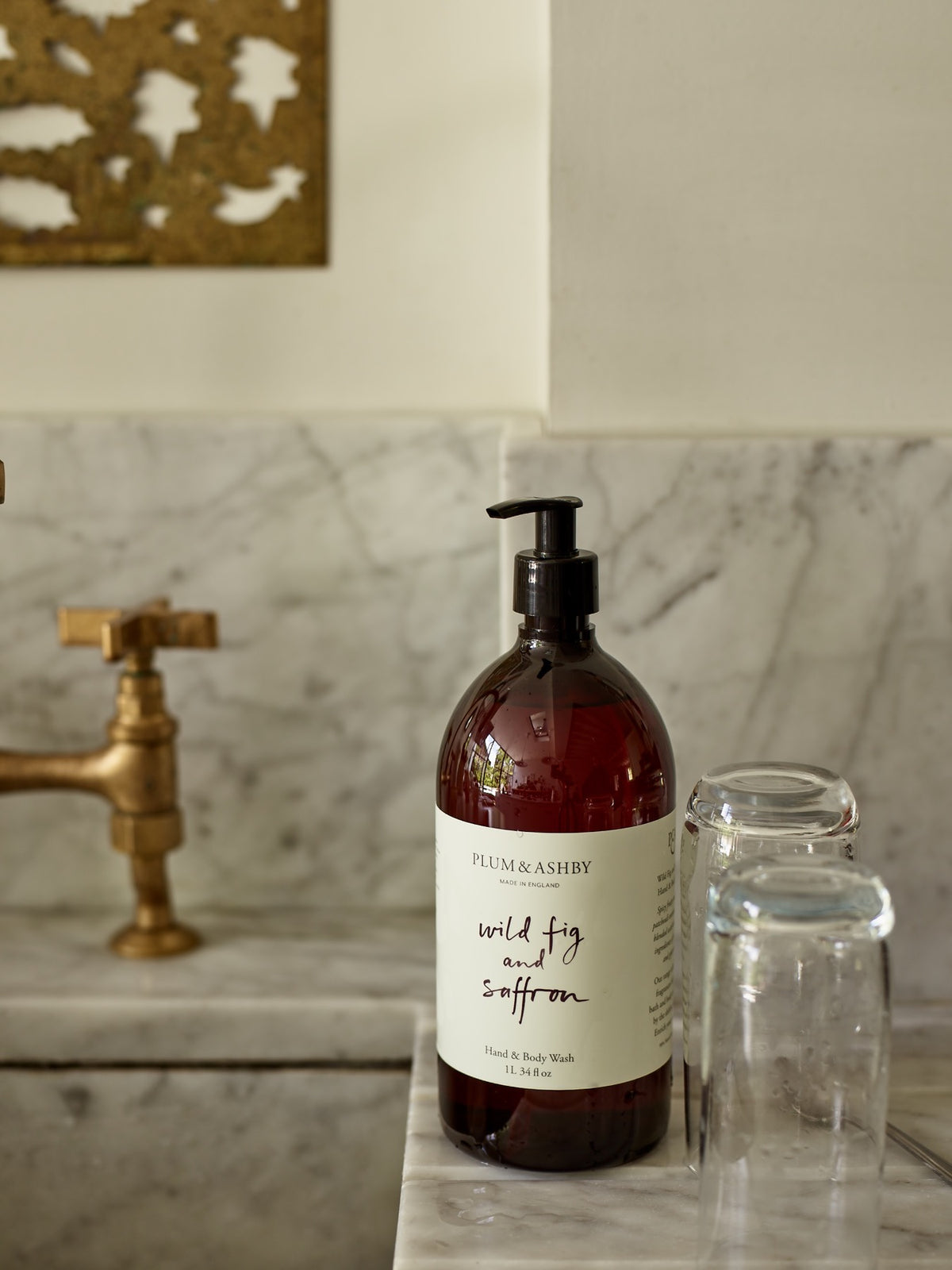 1 Litre bottle of Plum & Ashby Wild Fig & Saffron Hand & Body Wash on a kitchen countertop with a marble backdrop and a gold-patterned napkin holder in the background.