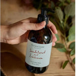 A pump bottle of Plum & Ashby Sandalwood and Labdanum hand and body wash, with a label that includes the product name and the words 'Made in England'. The bottle is held in a hand and there are plants in the background.