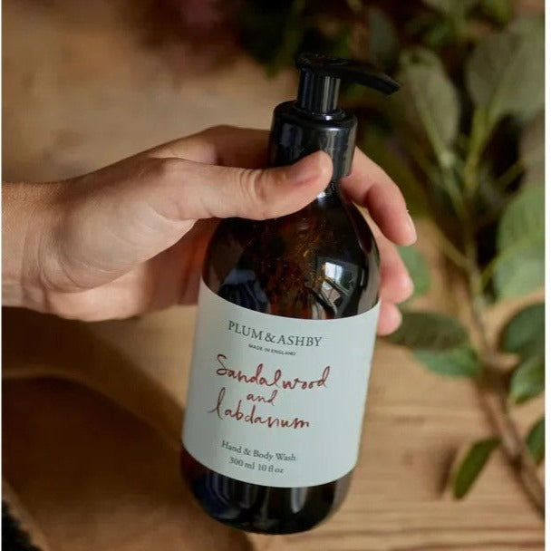 A pump bottle of Plum & Ashby Sandalwood and Labdanum hand and body wash, with a label that includes the product name and the words 'Made in England'. The bottle is held in a hand and there are plants in the background.