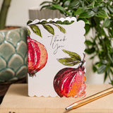 A scalloped white greeting card featuring watercolor illustrations of red pomegranates and green leaves, with the words 'Thank You' in black script. The card is displayed on a wooden surface with a gold pencil in the foreground and blurred potted plants in the background.