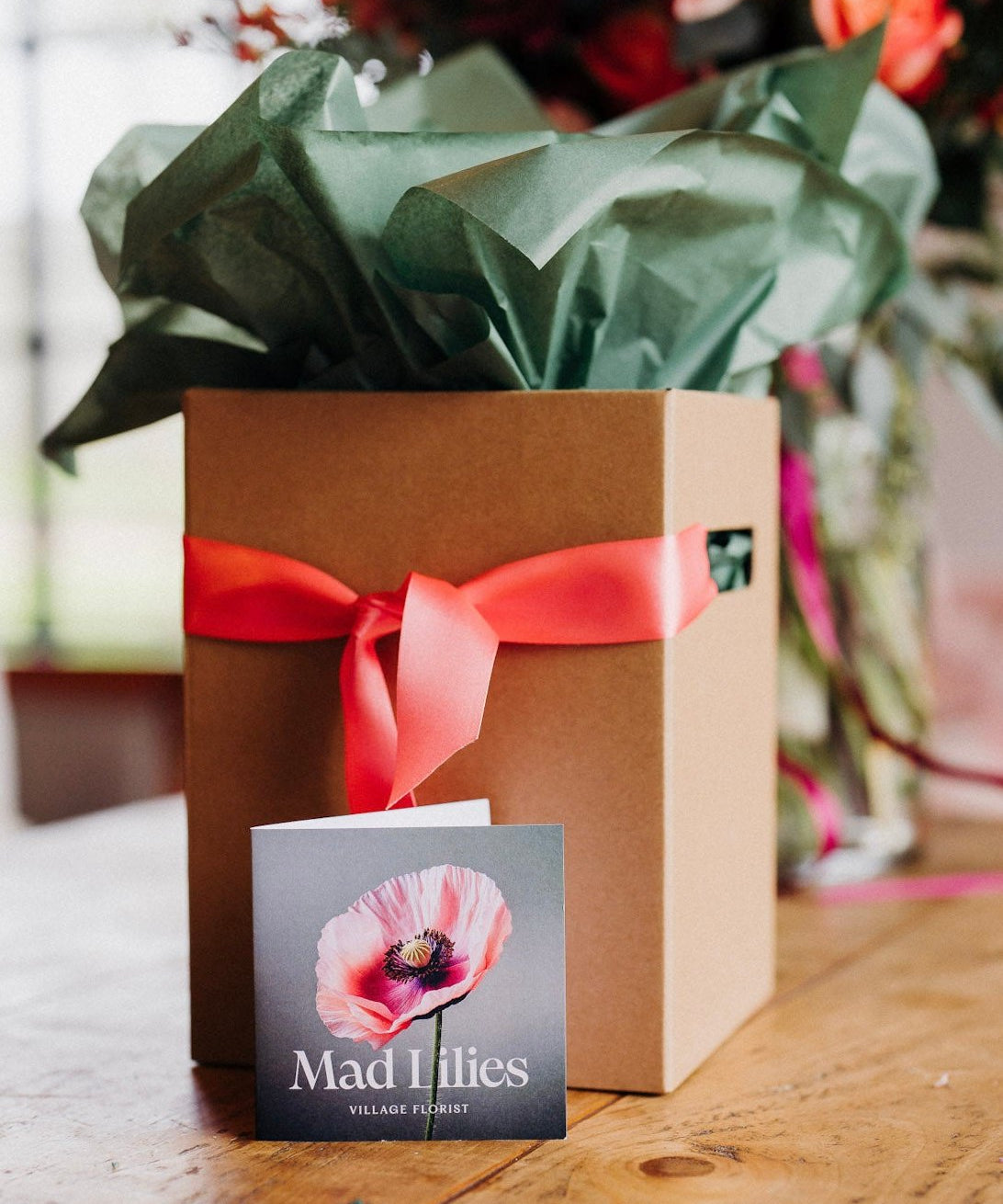 A kraft presentation box with moss green tissue paper and a coral satin ribbon, branded with a 'Mad Lilies' card in front.