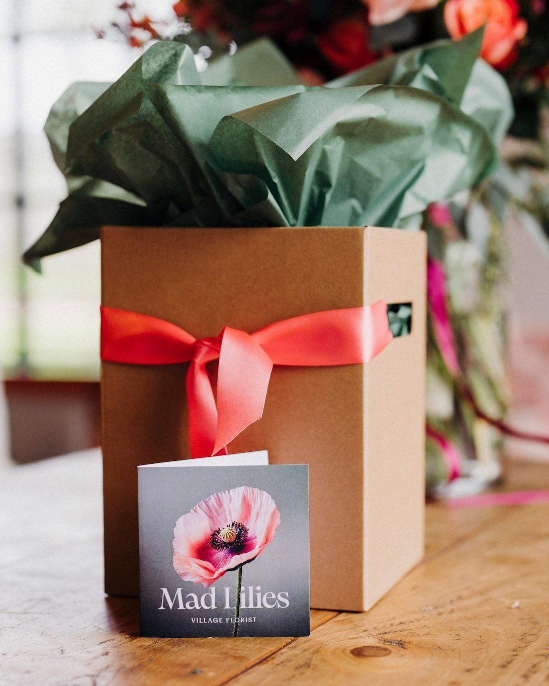 A kraft presentation box with moss green tissue paper and a coral satin ribbon, branded with a 'Mad Lilies' card in front.
