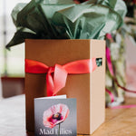 A kraft presentation box with moss green tissue paper and a coral satin ribbon, branded with a 'Mad Lilies' card in front.