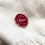 Red I Believe Christmas enamel pin badge with deep red enamel and metallic gold lettering on a backing card.