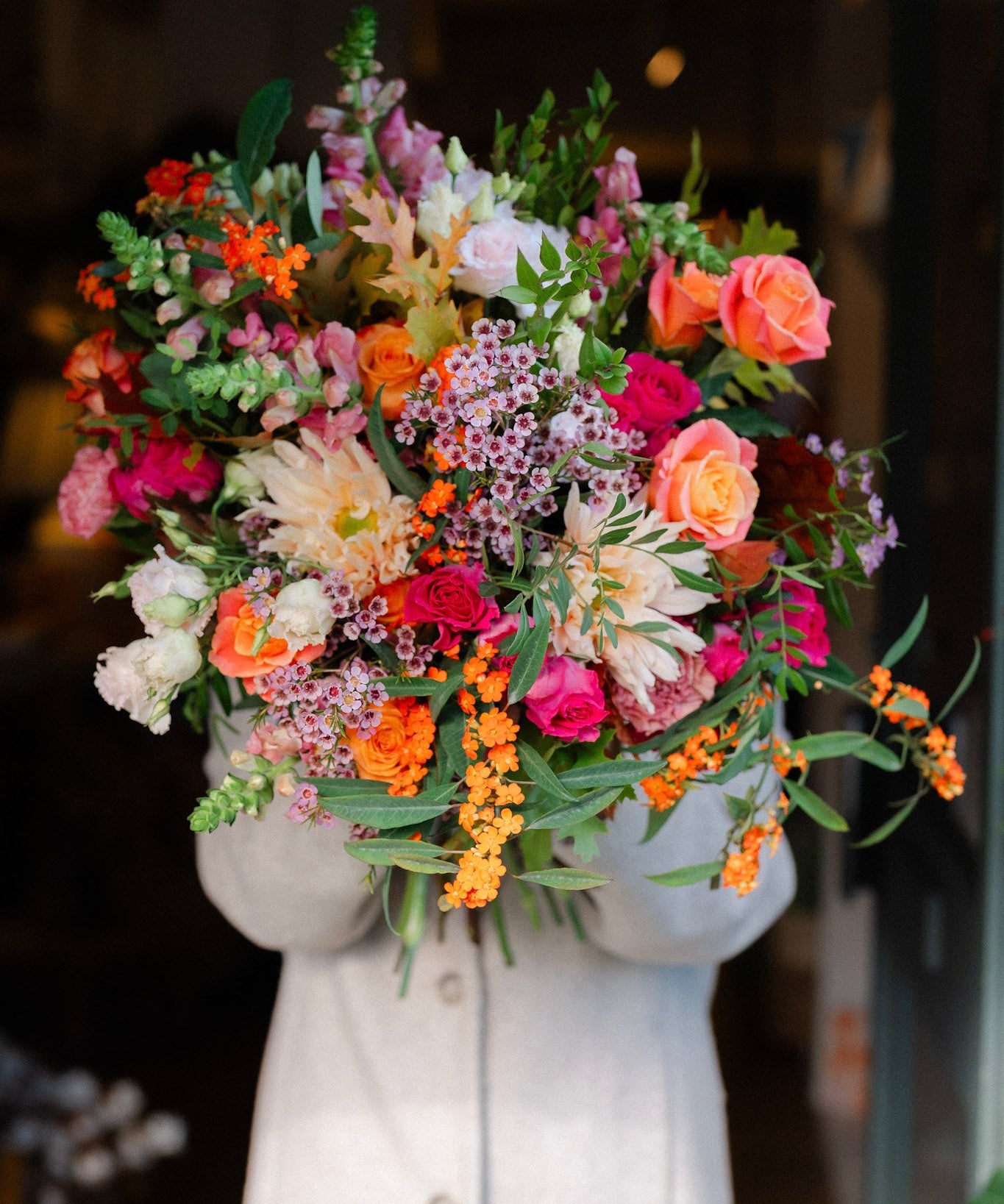 The Pink Bonfire autumn flower bouquet featuring orange Avignon blooms, apricot Miss Piggy roses, vibrant hot pink roses, waxflower and seasonal foliage.
