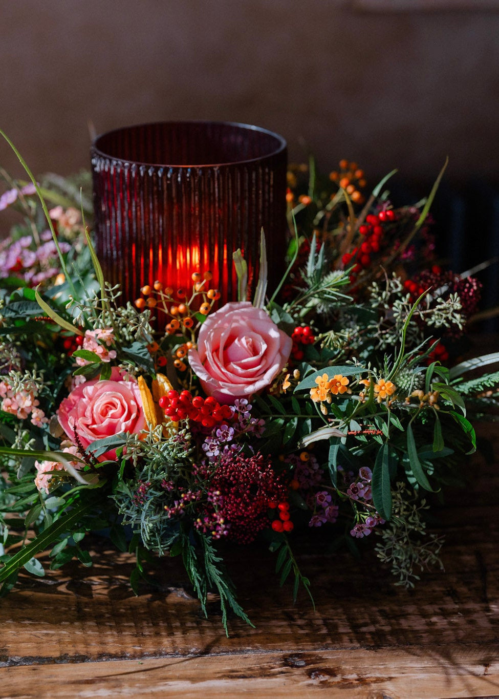 The Tipsy Truffle luxury Christmas floral table centrepiece with dusky pink roses, skimmia, berries, festive foliage and a ribbed glass hurricane candle holder