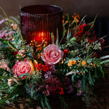 The Tipsy Truffle luxury Christmas floral table centrepiece with dusky pink roses, skimmia, berries, festive foliage and a ribbed glass hurricane candle holder