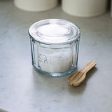 Vintage glass salt pot with lid and wooden spoon for kitchen storage.