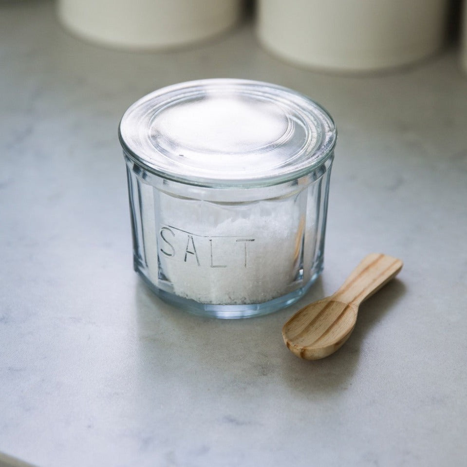 Vintage glass salt pot with lid and wooden spoon for kitchen storage.